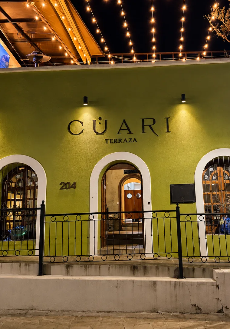 Fachada de CÜARI Terraza de noche, con el logotipo iluminado sobre la puerta y la terraza de la planta alta encendida.